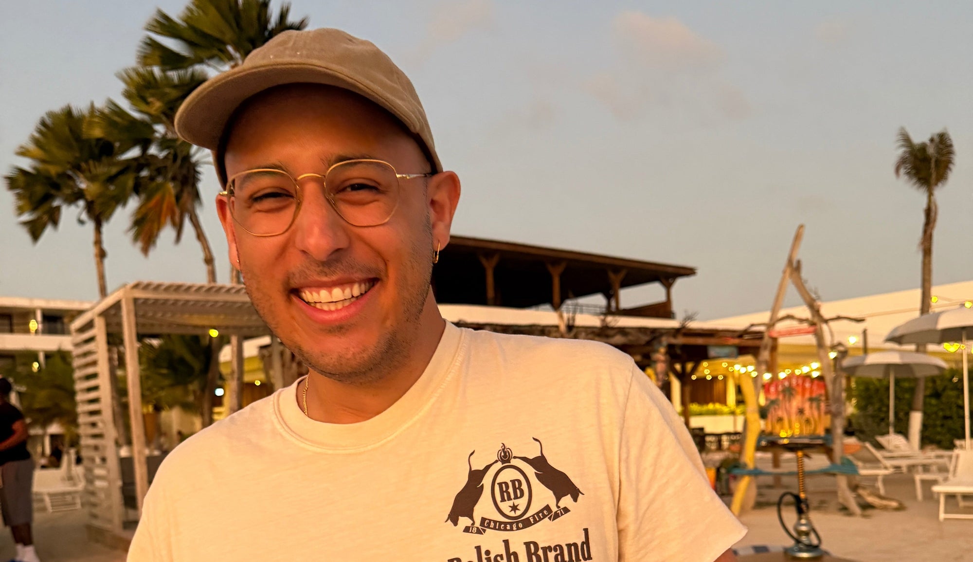 Man wearing a white t-shirt with a logo, holding a drink, standing on a beach with palm trees and a building in the background.