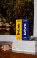 Two boxes of Teefpaste, one yellow and one blue, on a stack of books with a dark background.